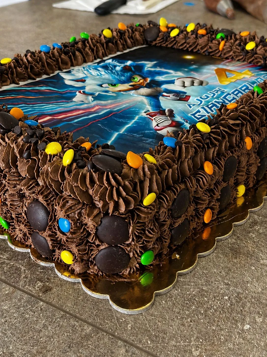 Inquadratura ravvicinata e laterale della torta di Sonic che mostra i dettagli dei ciuffi di ganache al cioccolato, i confetti Smarties gialli, blu e arancioni, e i grandi bottoni di cioccolato applicati sui lati.