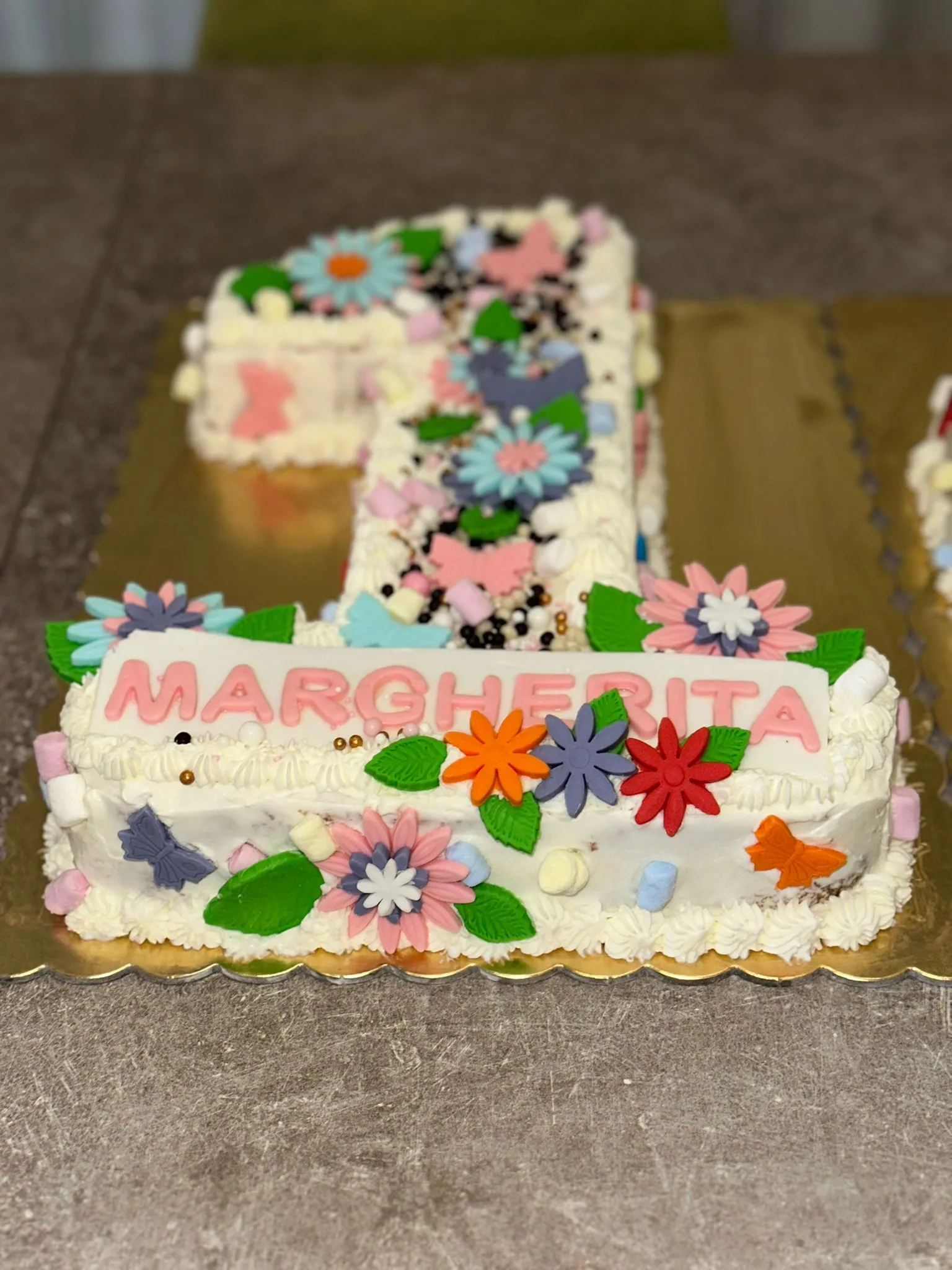 Inquadratura ravvicinata della torta a forma di numero uno ripiena con crema pasticcera e gocce di cioccolato, che mostra la targhetta in pasta di zucchero rosa con il nome Margherita, circondata da fiorellini, foglioline verdi e marshmallow.