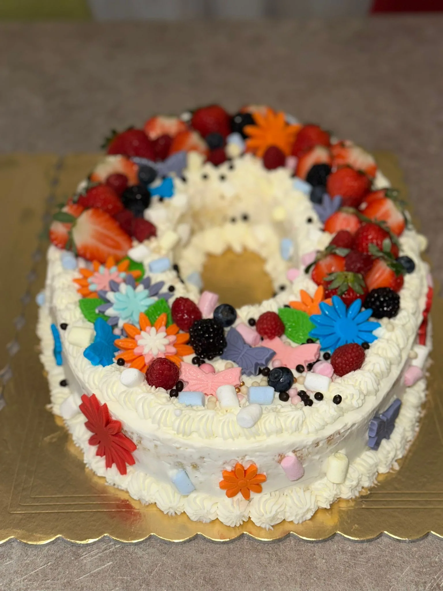 Inquadratura ravvicinata e angolata della torta a forma di numero zero ripiena di crema pasticcera e gragole, incentrata sulla ricca decorazione di fragole fresche, lamponi, mirtilli, marshmallow e fiorellini colorati.