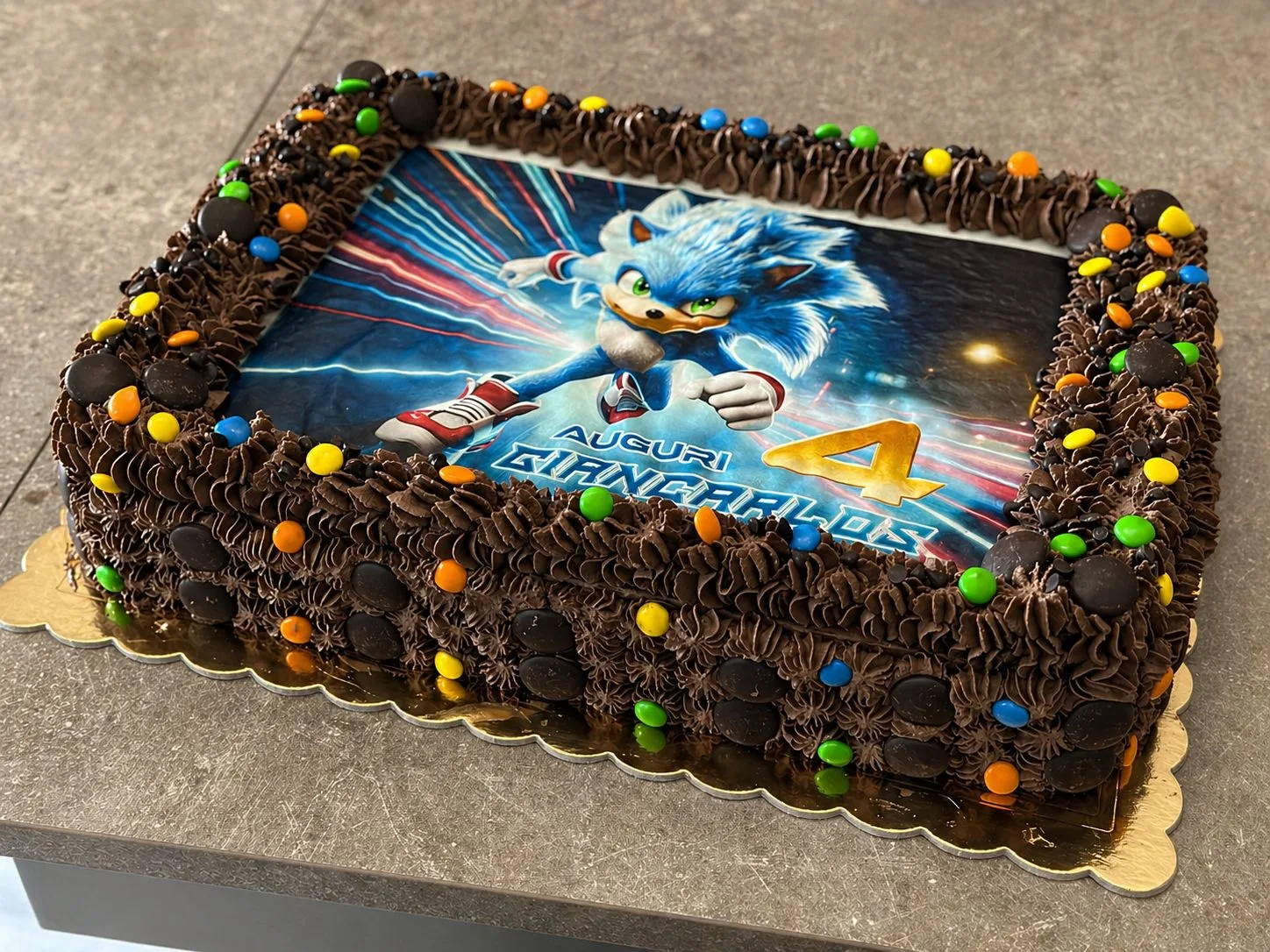 Vista panoramica dall'alto di una torta di compleanno rettangolare al cioccolato con cialda di Sonic, scritte di auguri per i 4 anni di Giancarlos e bordo decorato con Smarties colorati.