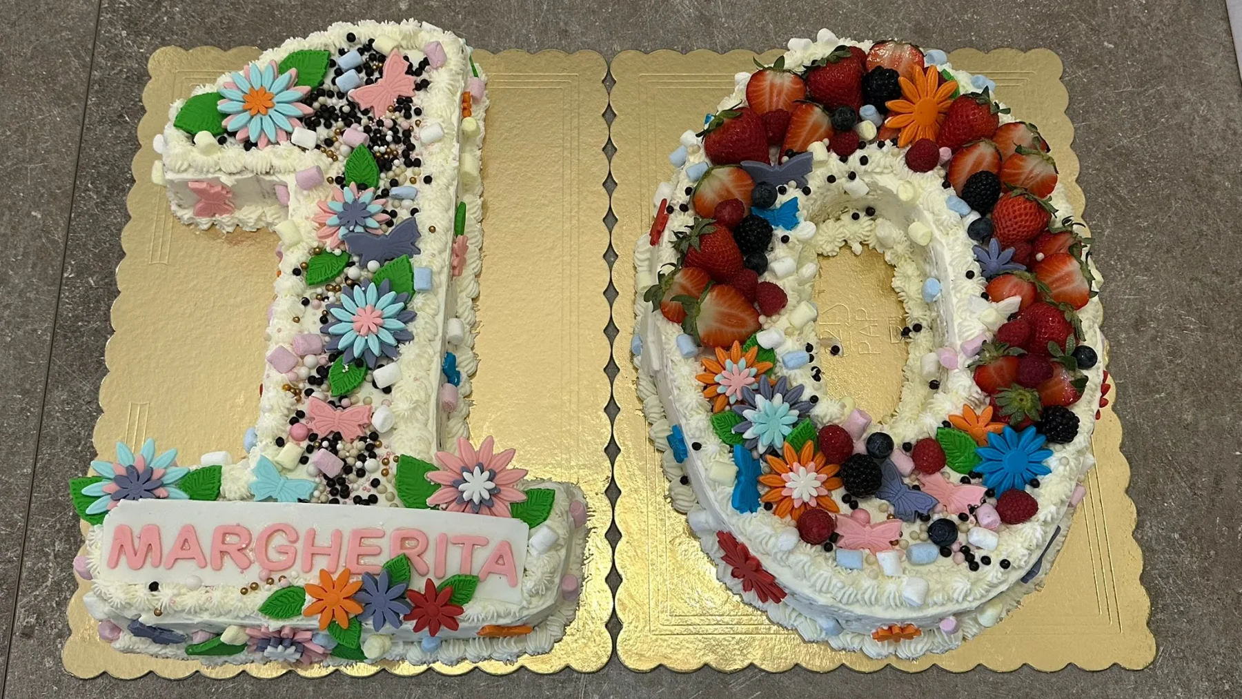Inquadratura frontale completa della torta di compleanno a forma di numero 10 ricoperta di panna, decorata con frutti di bosco, fiori in pasta di zucchero e la scritta Margherita sul numero uno.