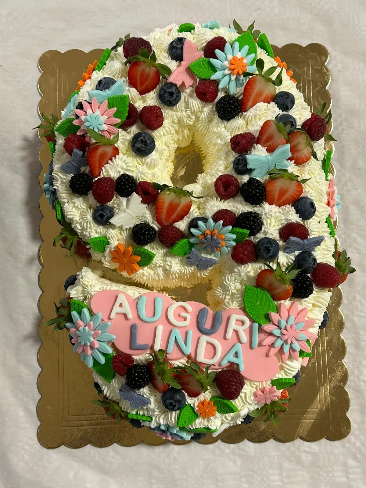Vista frontale completa della torta di compleanno a forma di numero 9 ricoperta di panna, con il nome Linda e decorazioni in pasta di zucchero a forma di farfalle e fiori rosa.