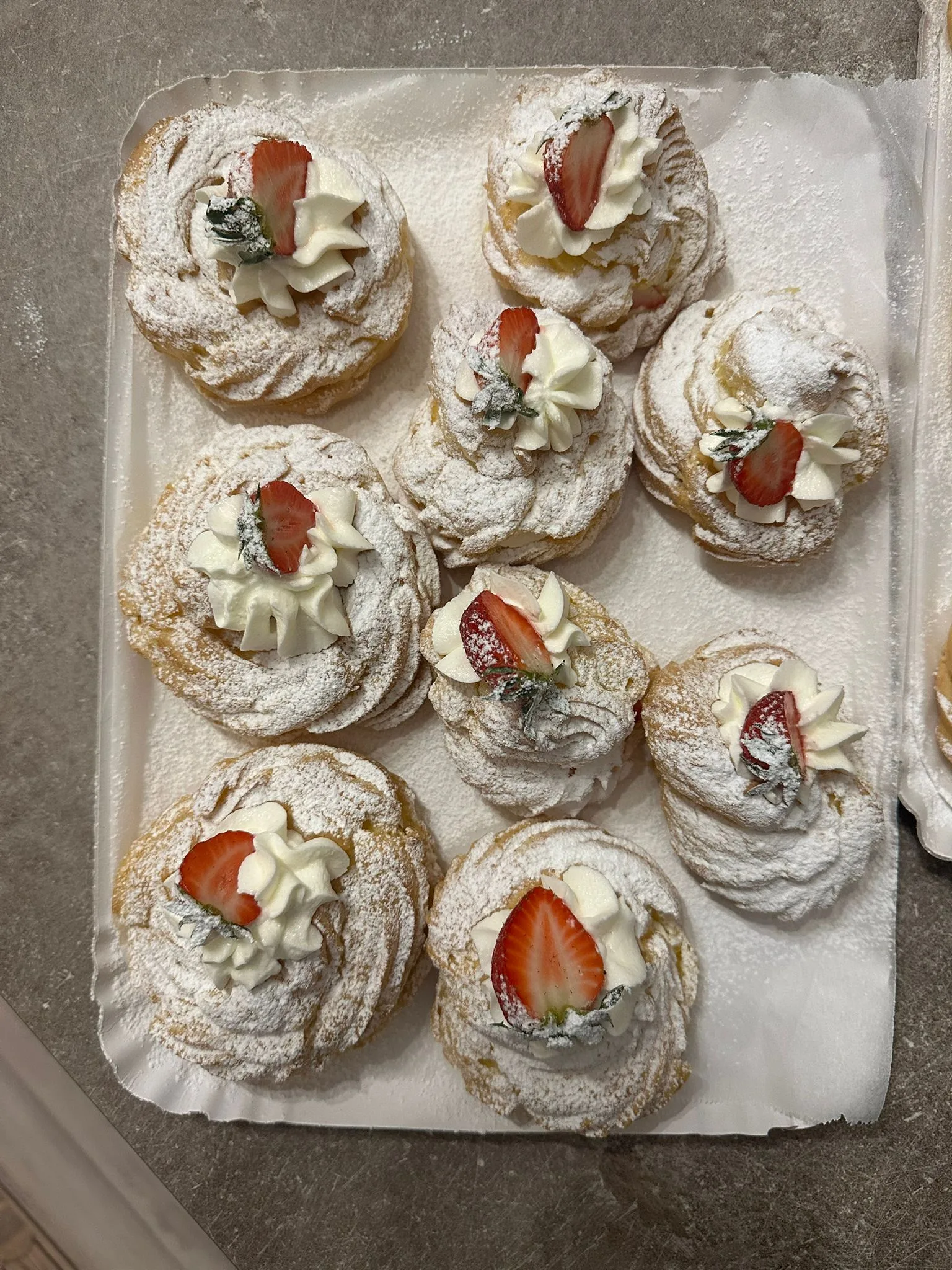 Inquadratura perpendicolare dall'alto di un vassoio con nove zeppole di San Giuseppe artigianali, ricoperte di zucchero a velo, ciuffi di panna e mascarpone e fette di fragola fresca al centro.