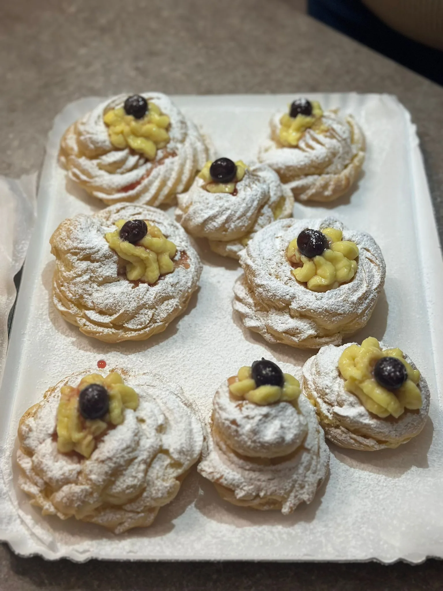 Inquadratura dall'alto di un vassoio bianco contenente otto zeppole di San Giuseppe artigianali, ricoperte di zucchero a velo e decorate singolarmente con ciuffi di crema pasticcera e un'amarena sciroppata.