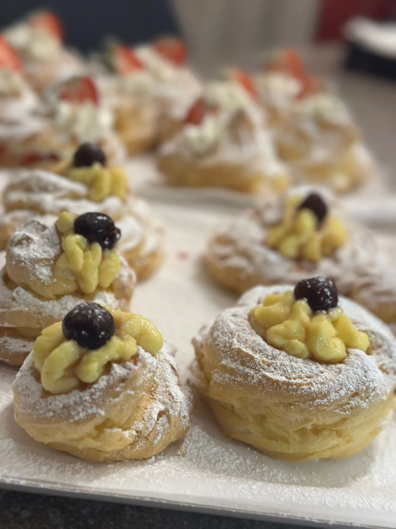 Inquadratura ravvicinata di un vassoio con Zeppole di San Giuseppe al forno in pasta choux, decorate con ciuffi di crema pasticcera gialla, zucchero a velo e un'amarena sciroppata al centro.