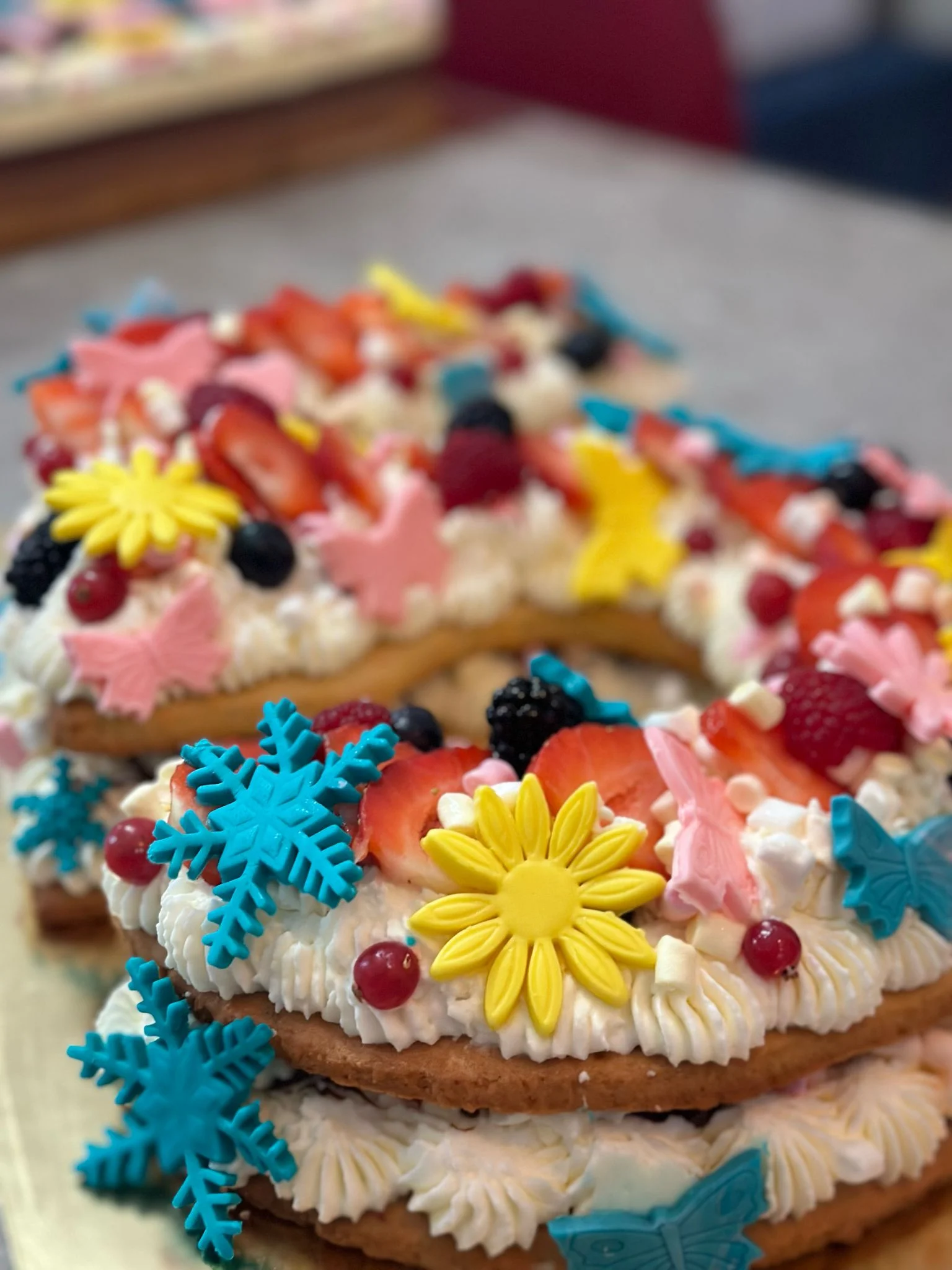 Dettaglio di una Cream Tart a forma di numero 5 a più strati, farcita con ciuffi di crema bianca e decorata con fragole, frutti di bosco, fiori gialli, farfalle rosa e fiocchi di neve azzurri.