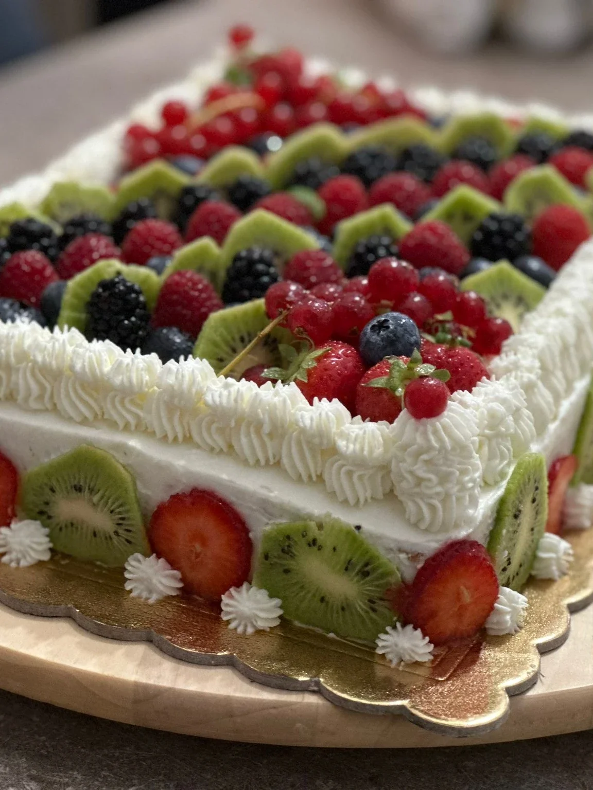 Grande torta rettangolare ricoperta di panna montata, decorata sulla superficie con file ordinate di kiwi, lamponi, more e ribes, e sui lati esterni con fette alternate di fragole e kiwi.